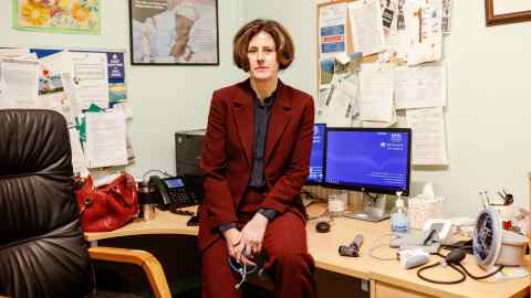 A woman in a dark red trouser suit sits on a desk in an office, holding a stethoscope