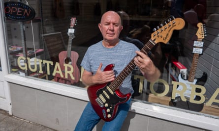Tony Galinos sits on the window sill outside his shop holding a guitar