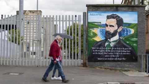 Two walkers with an umbrella pass a painted mural at the entrance to Casement Park